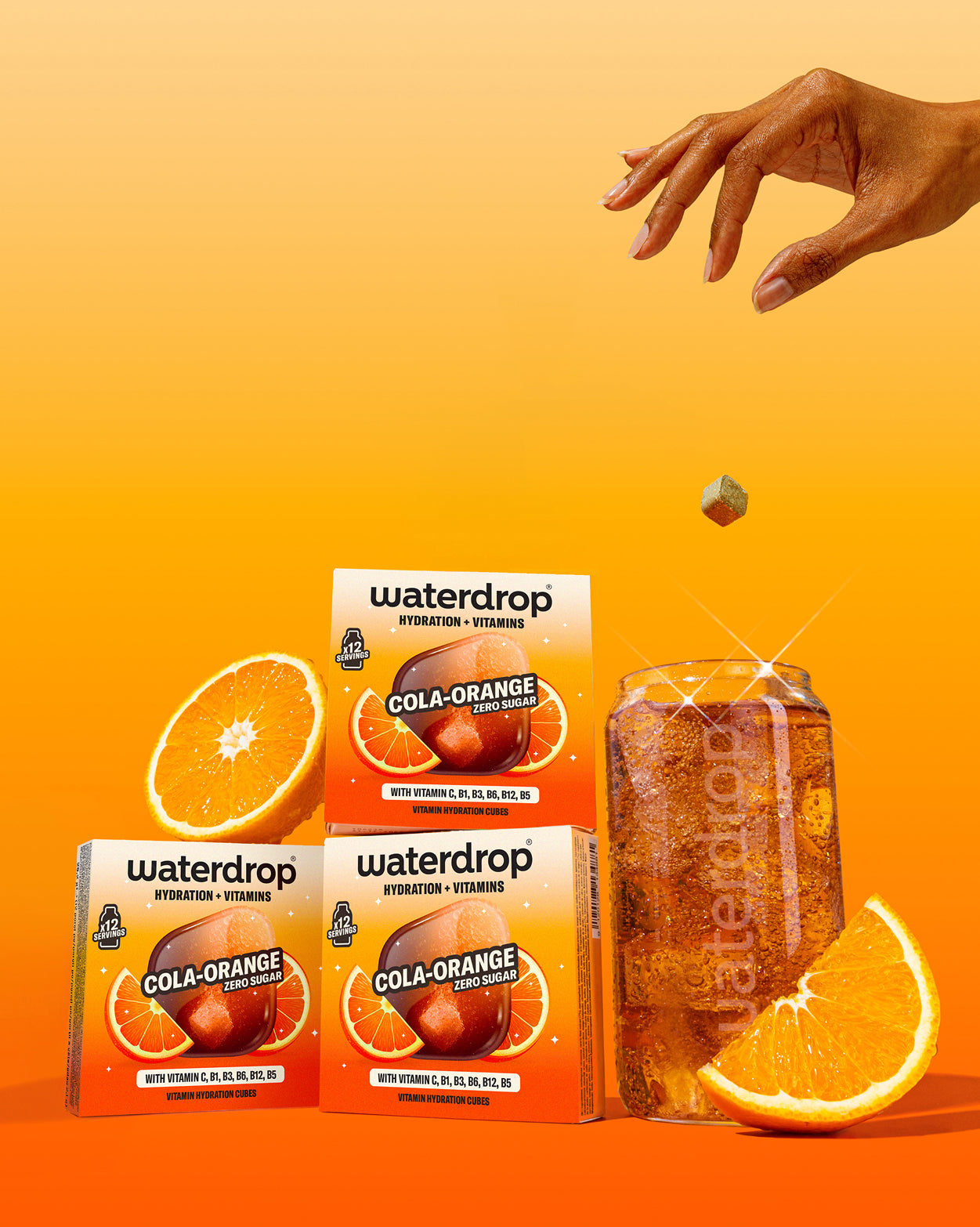 O mână lasă un cub Waterdrop Cola Orange să cadă într-un pahar cu băutură carbogazoasă, lângă trei cutii cu aroma Cola Orange și felii de portocală, pe fundal portocaliu.