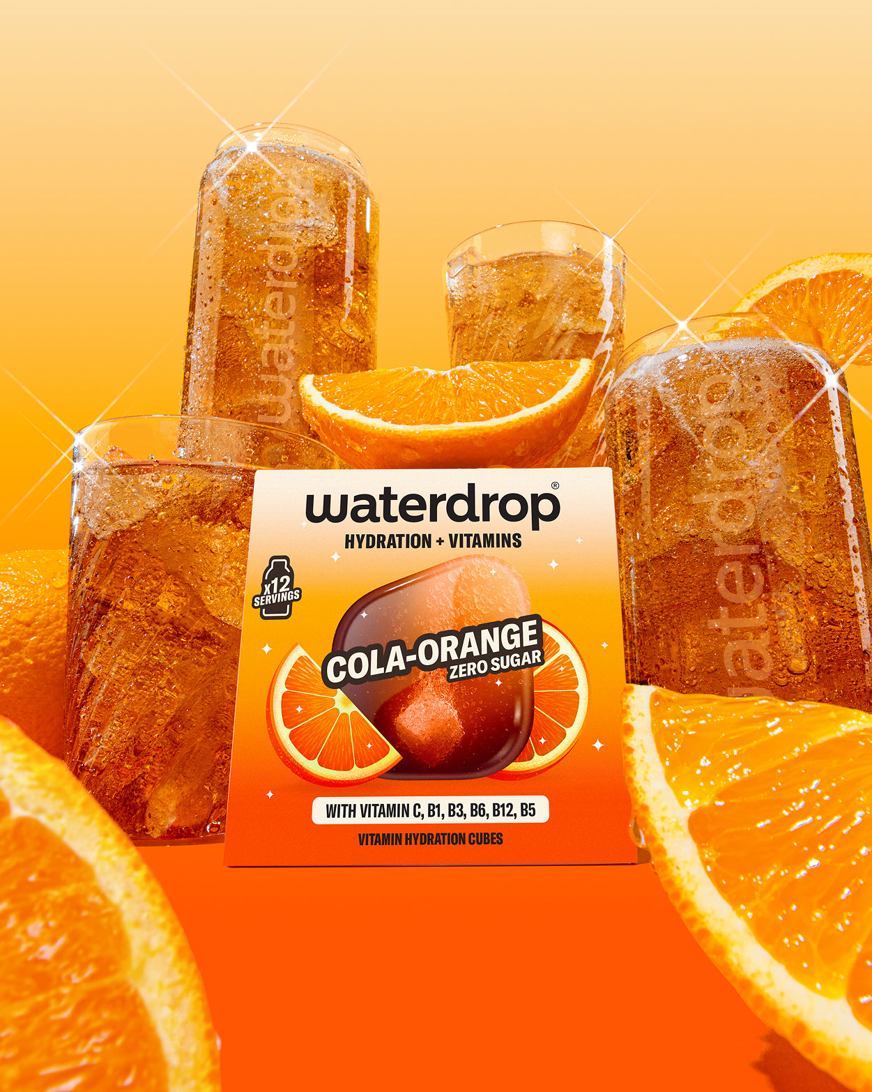 Pahare din sticlă Waterdrop umplute cu băutură carbogazoasă maro-portocalie, lângă cutia aromei Cola Orange și felii de portocală, pe fundal portocaliu.