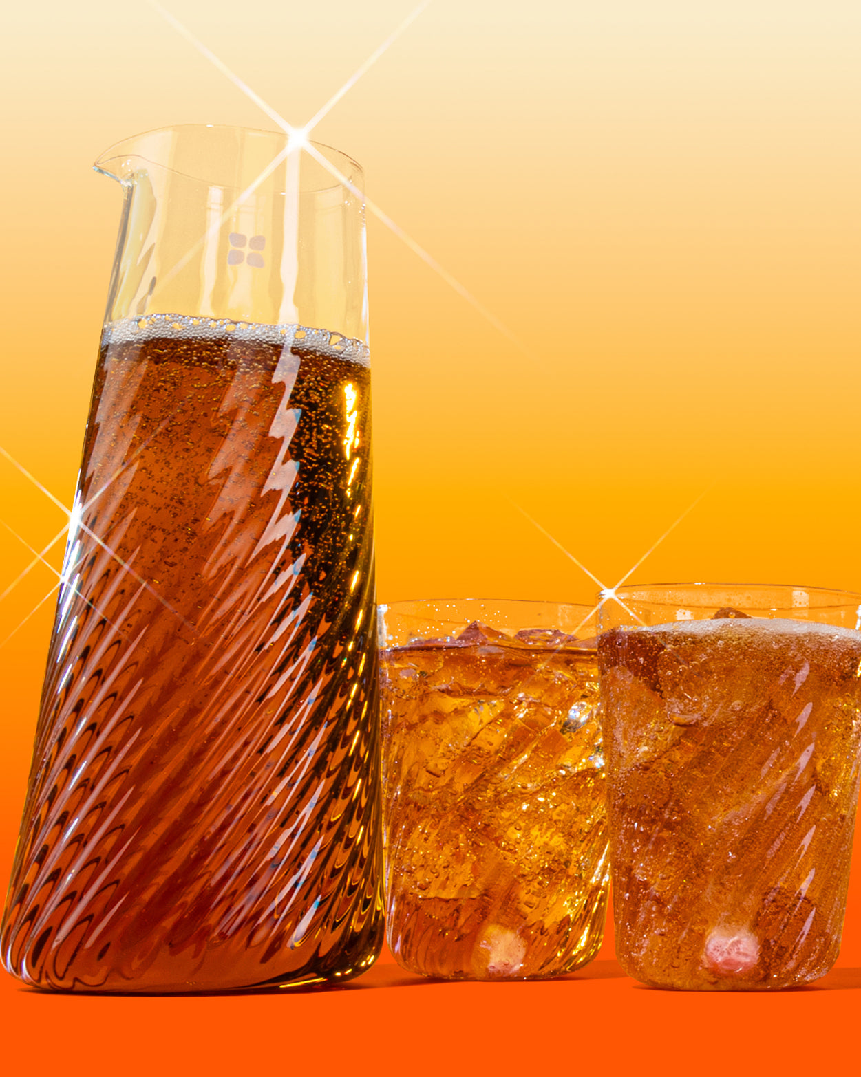Carafă din sticlă și două pahare spiralate umplute cu băutură carbogazoasă închisă la culoare cu gheață, pe fundal portocaliu.