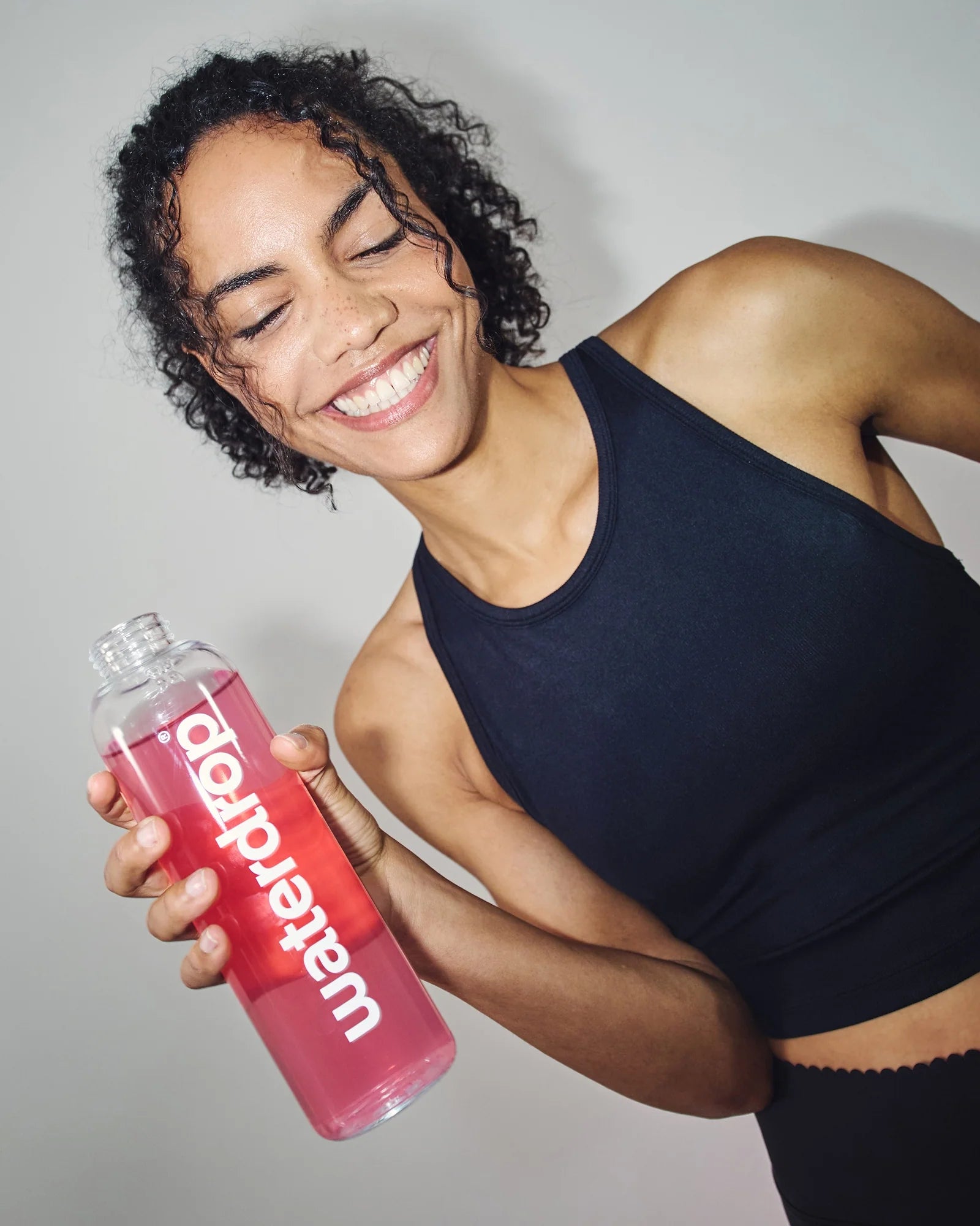 Femeie zâmbitoare îmbrăcată în echipament sport negru, ținând o sticlă waterdrop umplută cu băutură roz, pe fundal neutru.