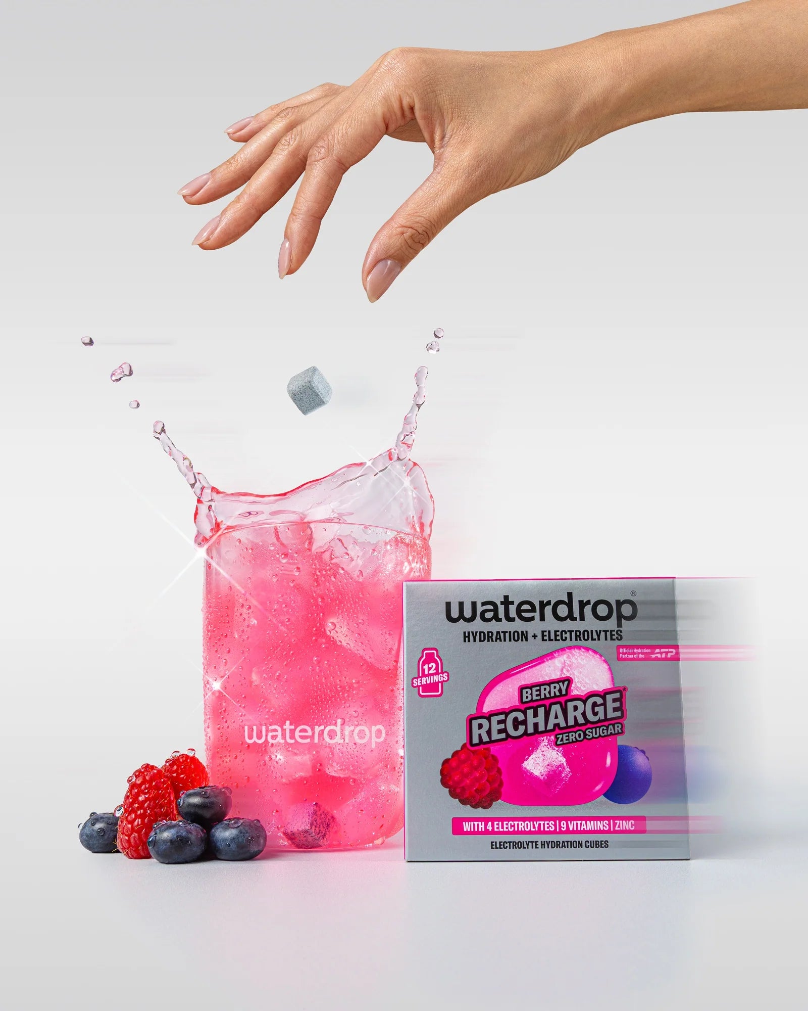 Mână deasupra unui pahar waterdrop cu băutură roz și cuburi de gheață, cu un cub de hidratare care cade în pahar, lângă o cutie Berry Recharge fără zahăr cu 12 porții și fructe de pădure.