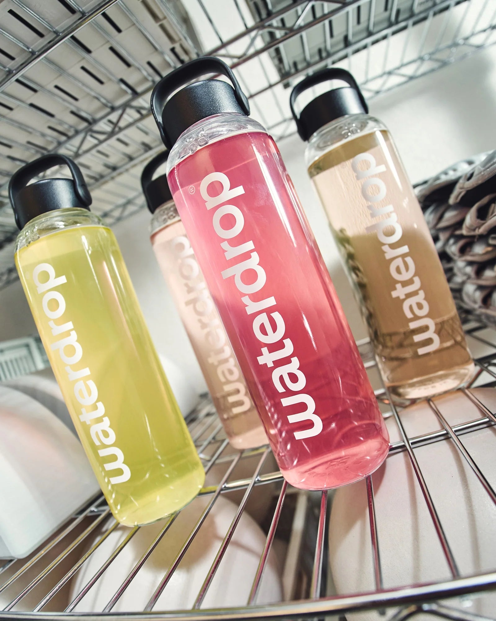 Patru sticle reutilizabile waterdrop așezate pe un raft metalic, umplute cu băuturi în culori diferite (galben, roz, bej și maro deschis), fiecare cu capac negru și mâner.