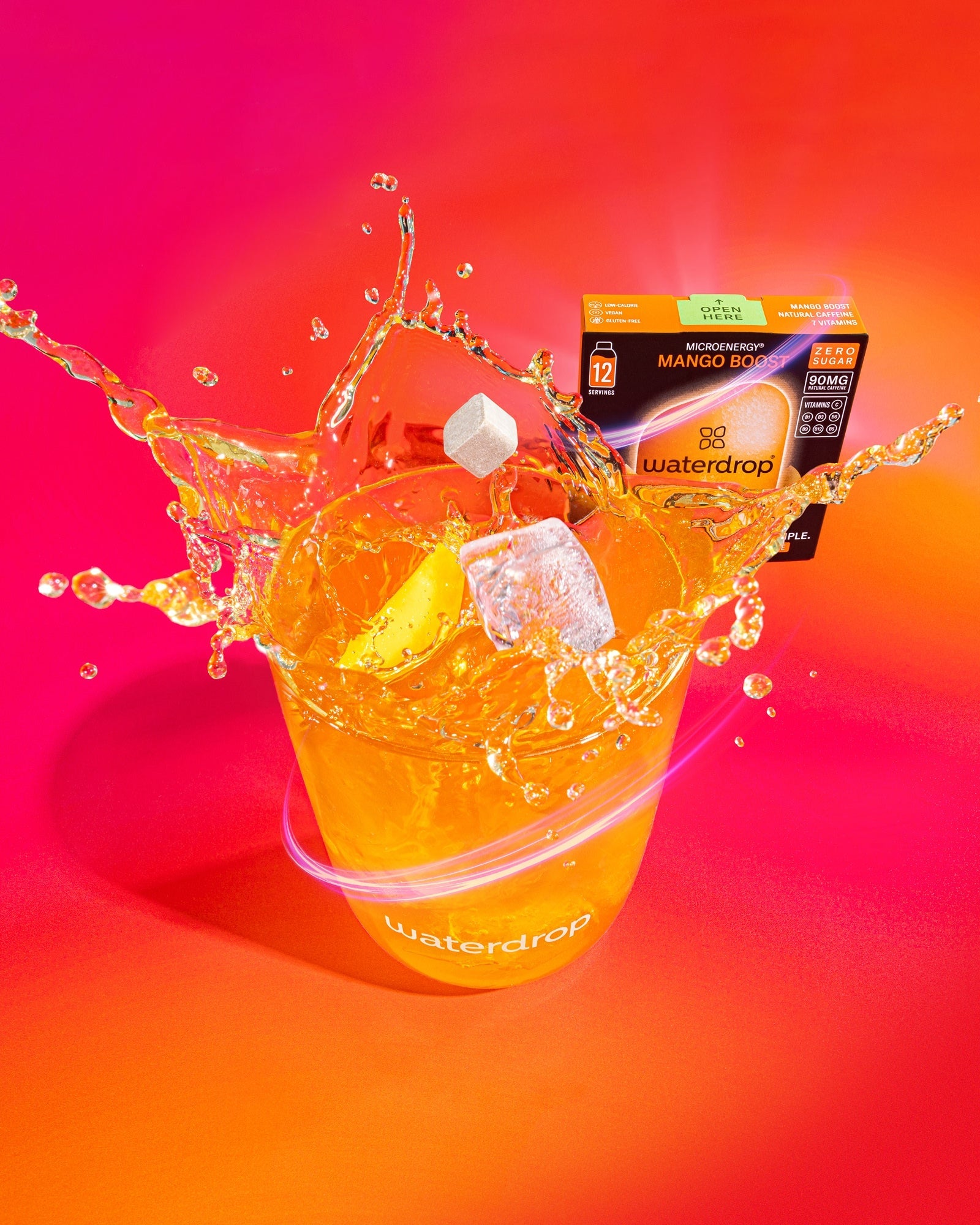 Pahar waterdrop cu băutură Mango Boost, cuburi de gheață și stropi de lichid, cu o cutie Microenergy Mango Boost fără zahăr cu 12 porții în fundal.