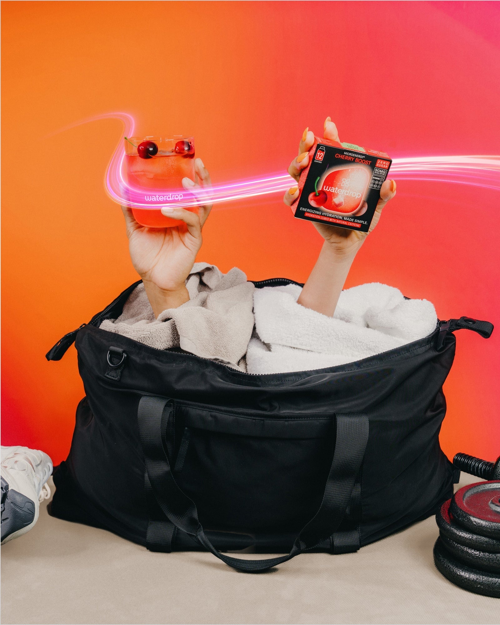 Mâini care ies dintr-o geantă sport neagră, ținând un pahar waterdrop cu băutură roșie și cireșe și o cutie Microenergy Cherry Boost fără zahăr cu 12 porții, pe fundal portocaliu.
