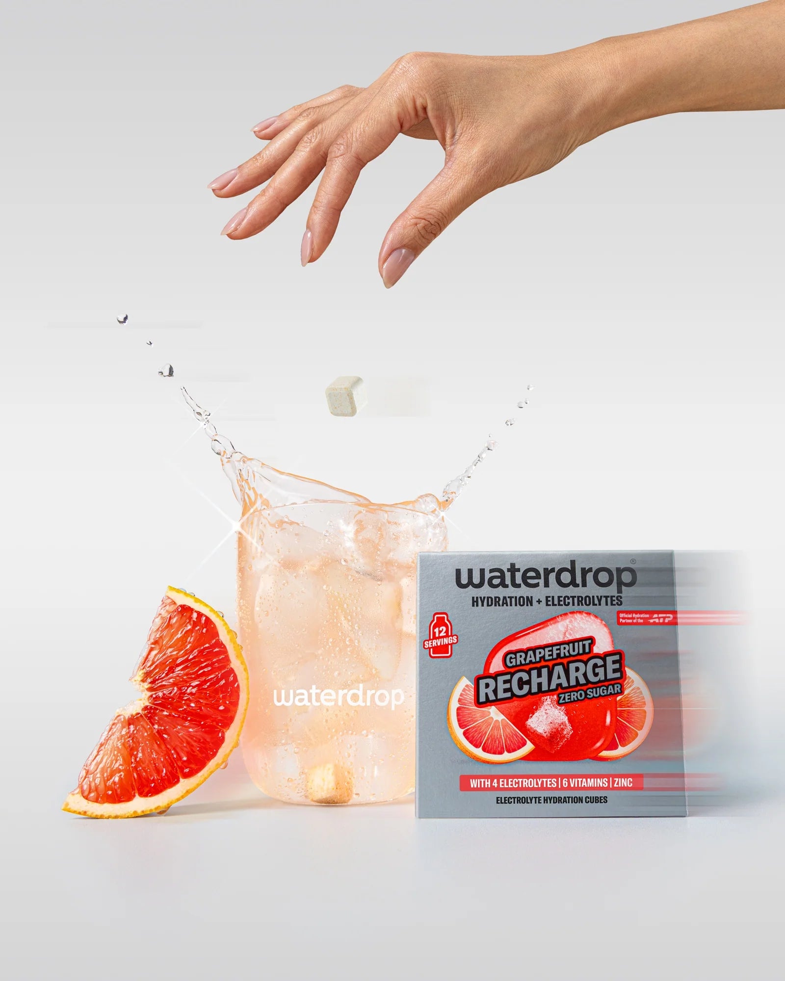 Mână deasupra unui pahar waterdrop cu băutură grapefruit și cuburi de gheață, cu un cub de hidratare care cade în pahar, lângă o cutie Grapefruit Recharge fără zahăr cu 12 porții și o felie de grapefruit.