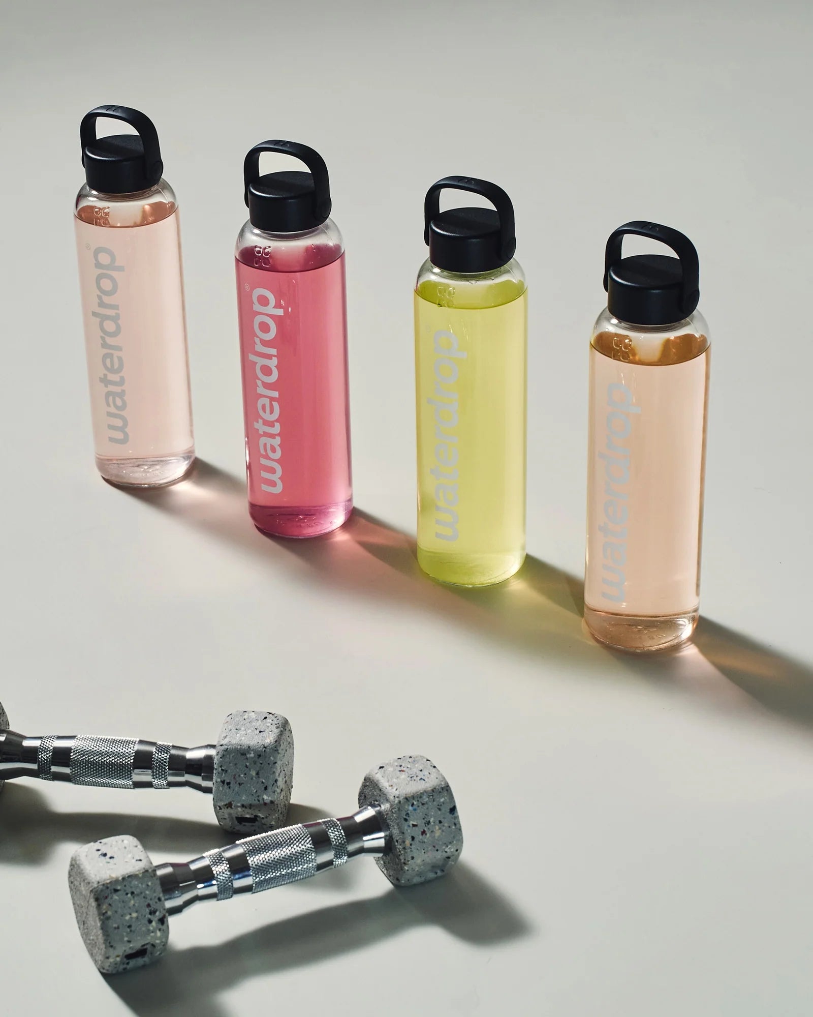 Patru sticle reutilizabile waterdrop aliniate, umplute cu băuturi în nuanțe diferite (roz deschis, roz, galben și piersică), cu gantere metalice în prim-plan, pe fundal deschis.