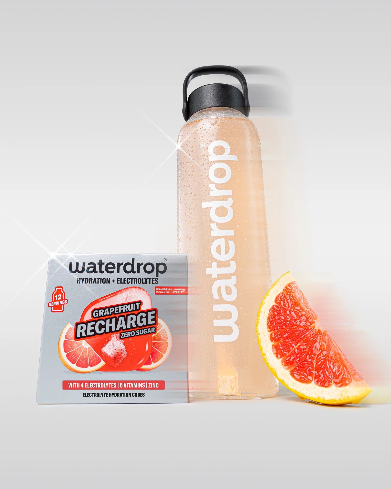 Sticlă reutilizabilă waterdrop umplută cu băutură roz grapefruit, lângă o cutie Grapefruit Recharge fără zahăr cu 12 porții și o felie de grapefruit, pe fundal alb.