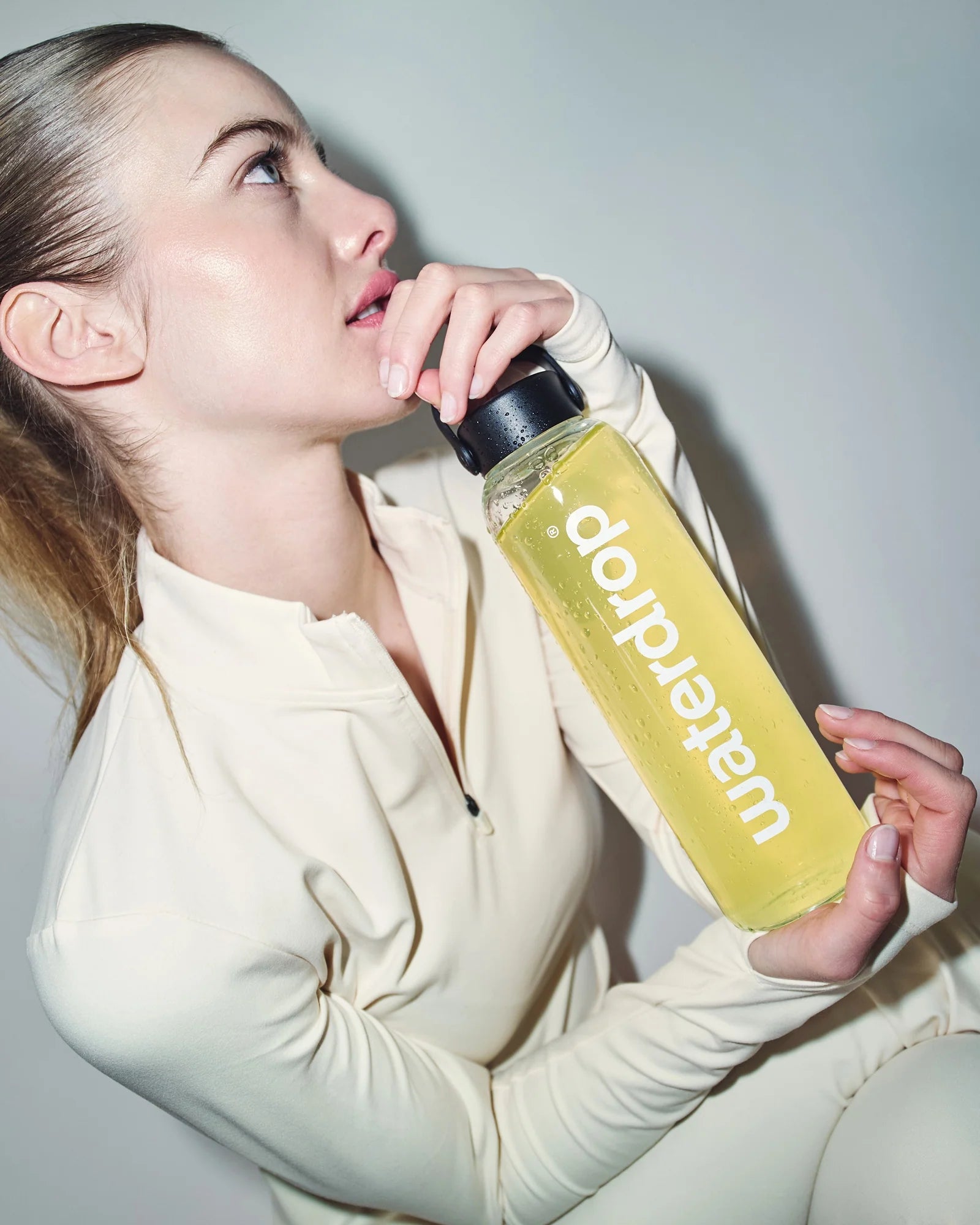 Femeie în echipament sport deschis la culoare, ținând o sticlă waterdrop umplută cu băutură galbenă Lemon-Mint, pe fundal neutru.