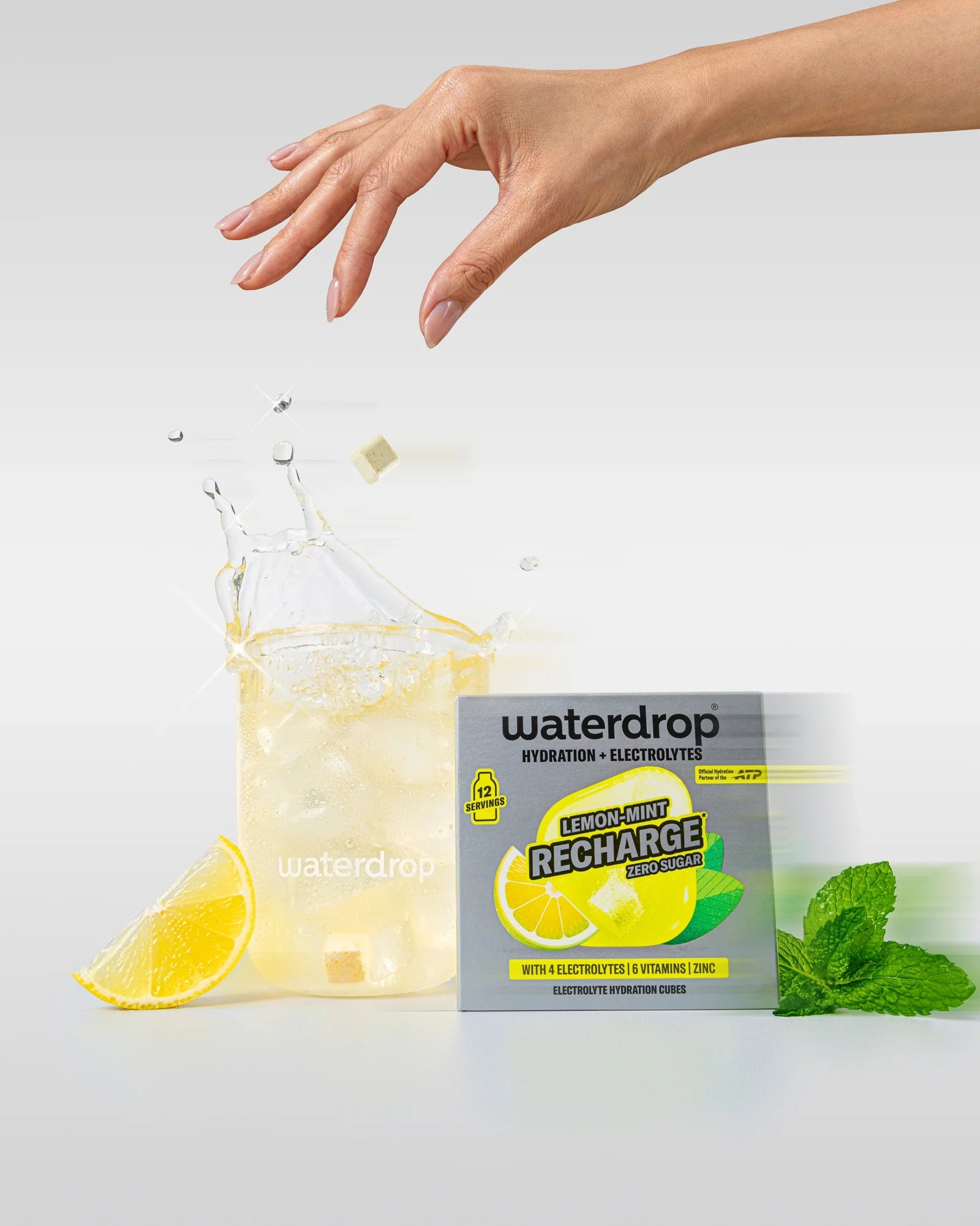 Mână deasupra unui pahar waterdrop cu băutură Lemon-Mint și cuburi de gheață, cu un cub de hidratare care cade în pahar, lângă o cutie Lemon-Mint Recharge fără zahăr cu 12 porții, felie de lămâie și mentă.