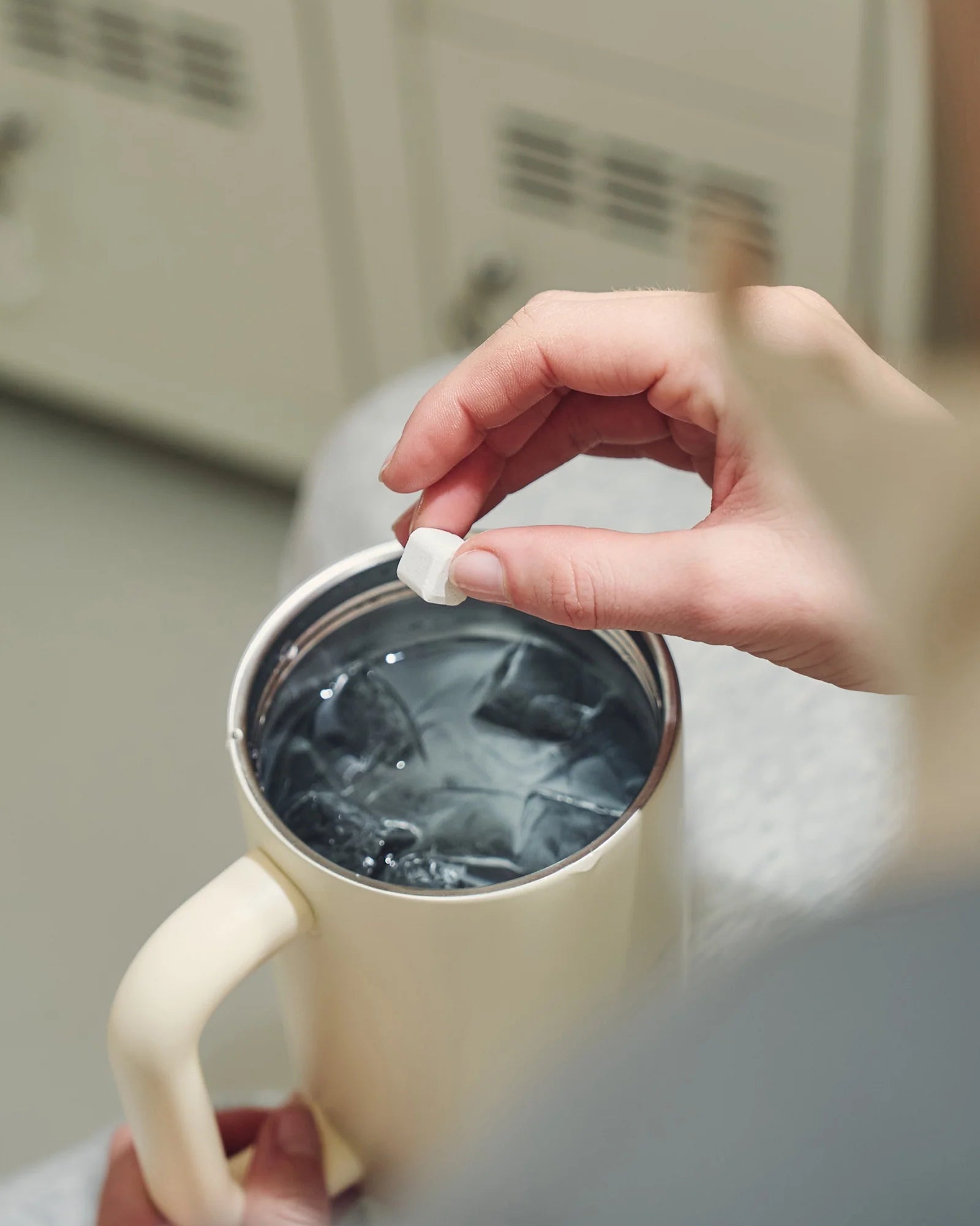 Mână care ține un cub de hidratare deasupra unei căni termos bej waterdrop umplute cu apă și gheață, văzută de sus.