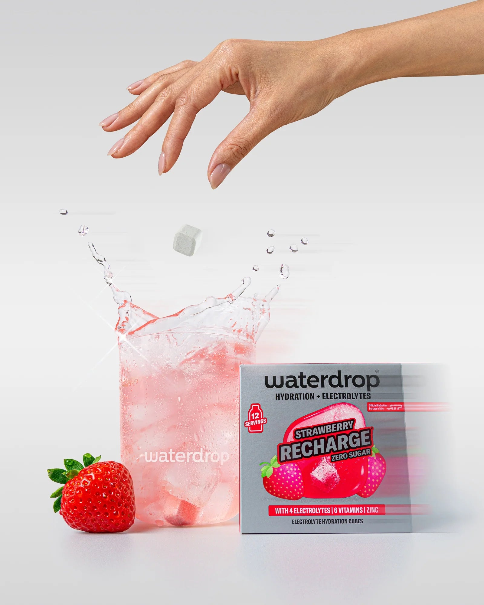 Mână deasupra unui pahar waterdrop cu băutură roz de căpșuni și cuburi de gheață, cu un cub de hidratare care cade în pahar, lângă o cutie Strawberry Recharge fără zahăr cu 12 porții.