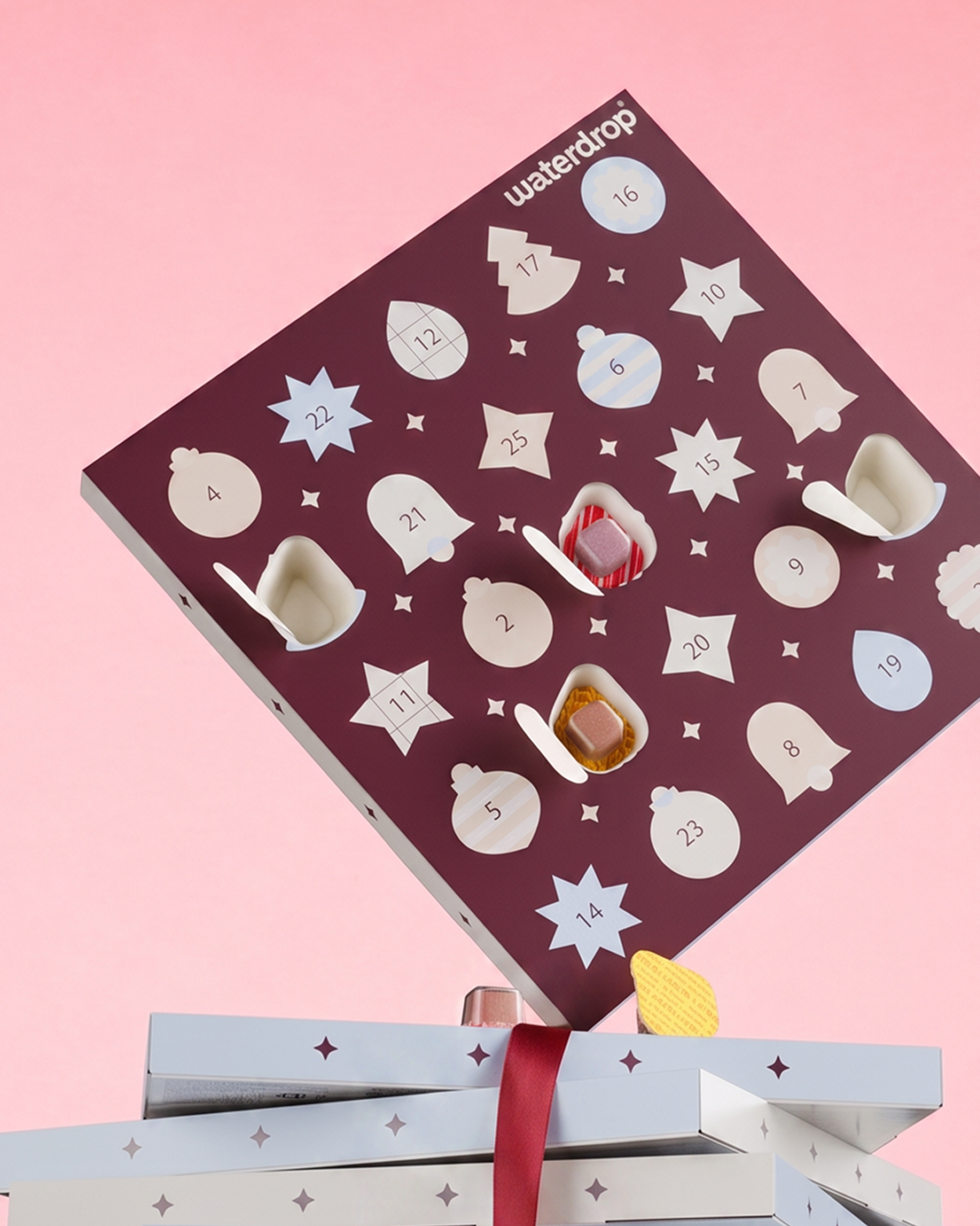 Calendar de Advent mov cu uși în formă de stele numerotate pe fond roz, plasat deasupra a două cadouri împachetate.