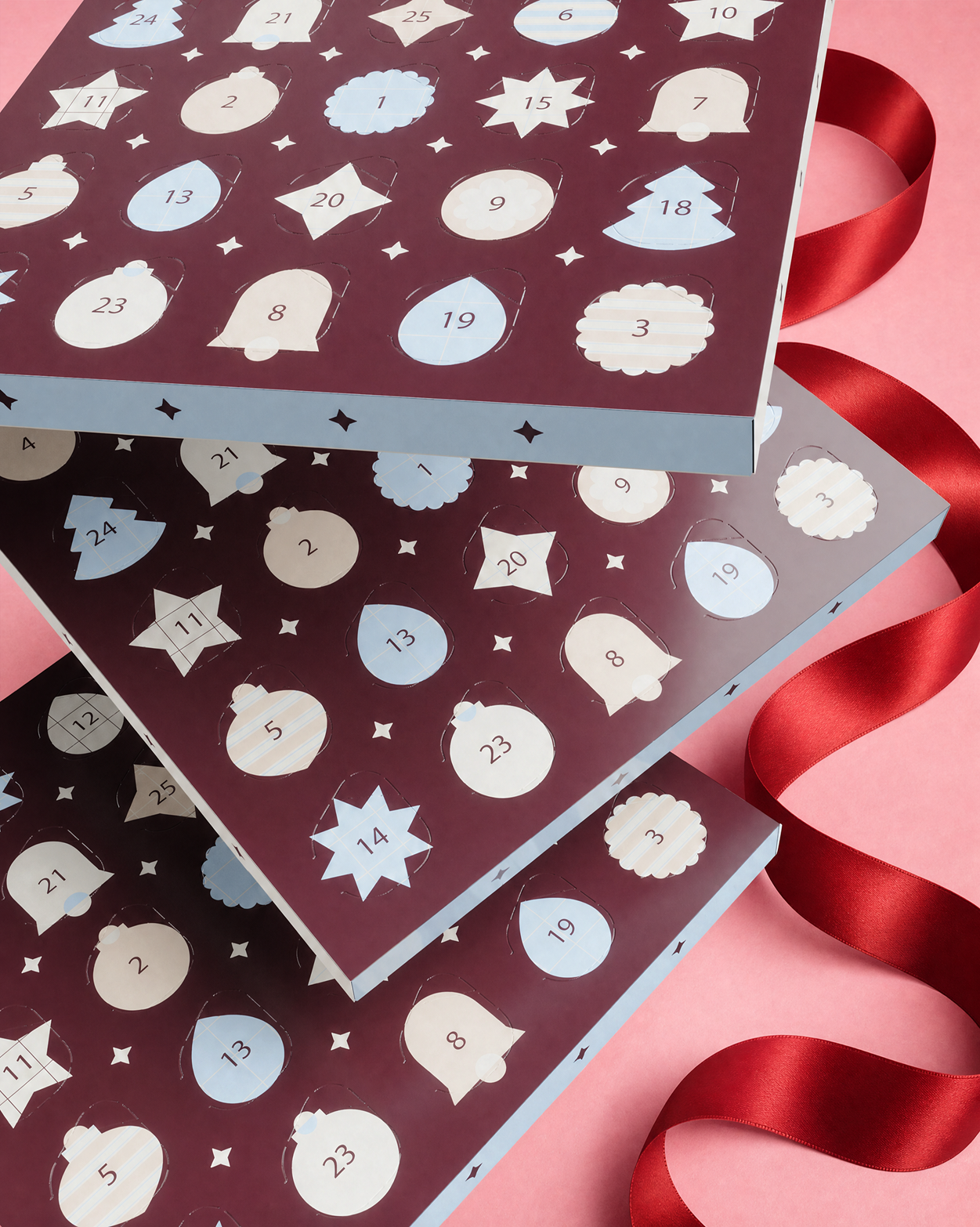 Trei cutii de Advent suprapuse cu uși numerotate și forme decorative, legate cu o panglică roșie pe fundal roz.