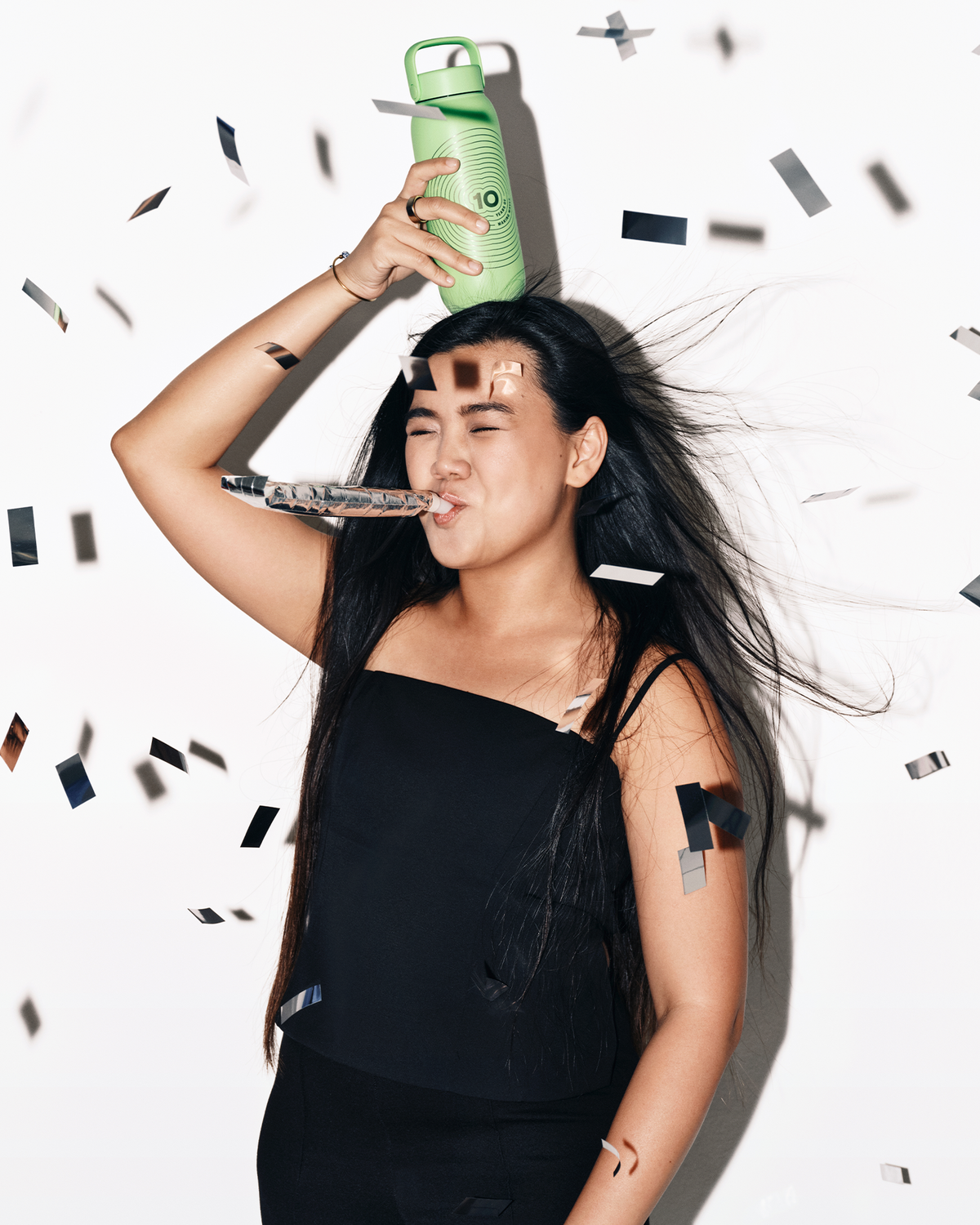Femeie într-o bluză neagră ține o sticlă de spray verde deasupra capului, în jur cad confetti.