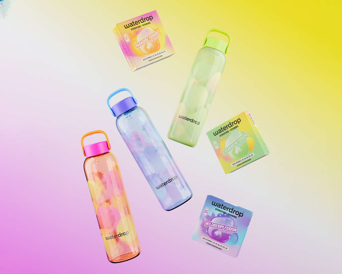 Set de patru sticle de apă transparente cu dopuri colorate, așezate pe un fundal gradient pastel, alături de ambalajele lor.