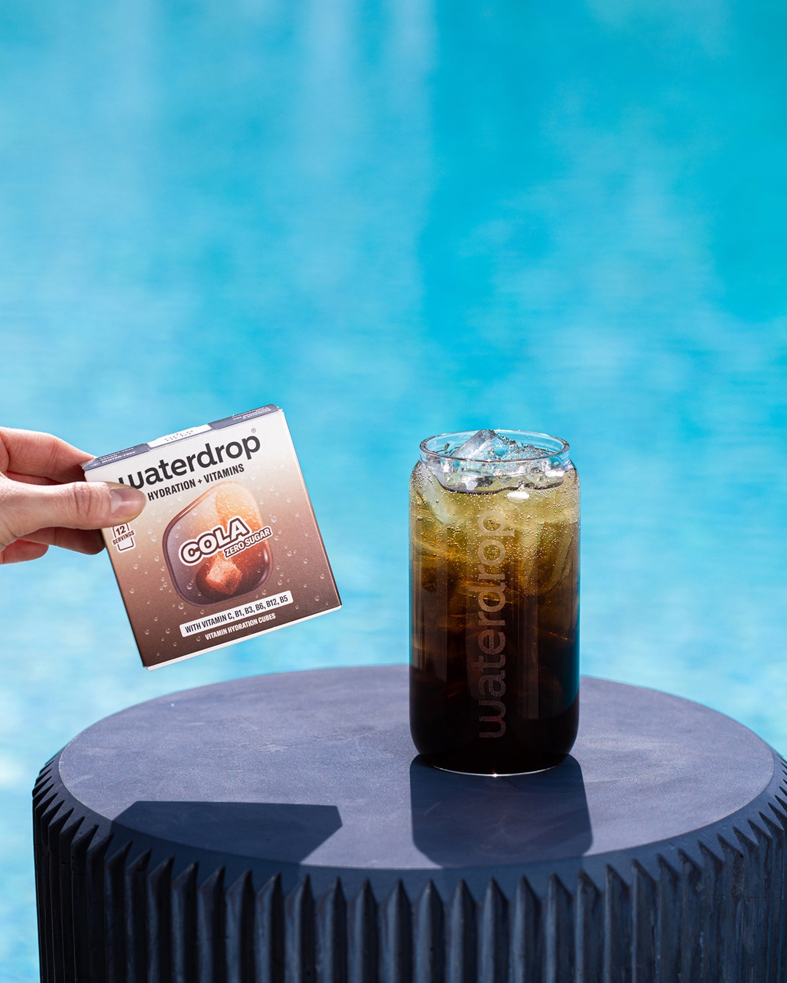 O mână ține un pliculeț lângă un pahar cu cola rece pe un piedestal albastru rotund în apropierea piscinei.